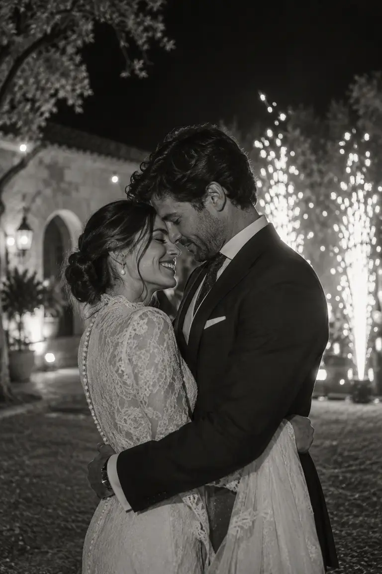 Retrato en blanco y negro de una pareja abrazada, con chispas frías y una casa de piedra desenfocadas al fondo.